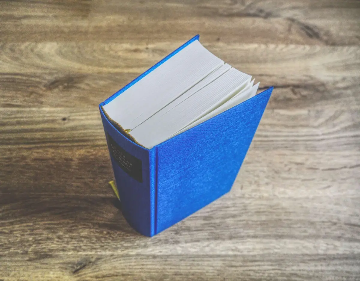 Ein dickes, gebundenes Buch mit blauem Einband liegt aufgeschlagen auf einer hölzernen Unterlage und gibt den Blick auf weiße Seiten und ein Lesezeichen mit goldenem Band frei.