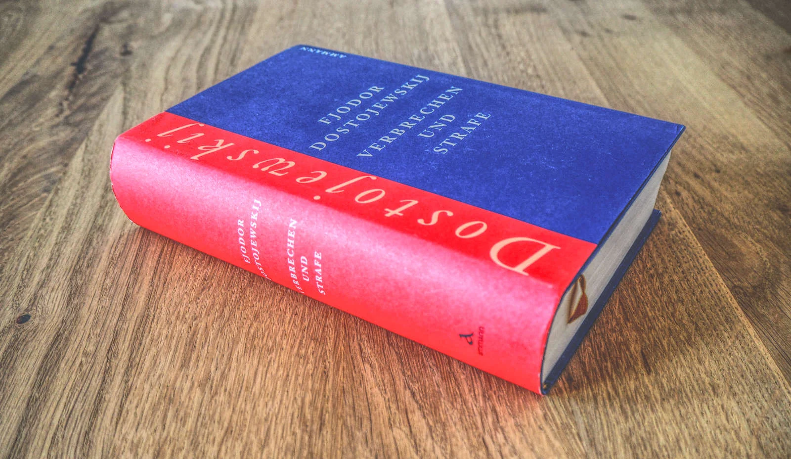 Auf einer Holzfläche liegt ein gebundenes Exemplar von Fjodor Dostojewskis „Verbrechen und Strafe“. Der Einband ist überwiegend blau, der Buchrücken rot.