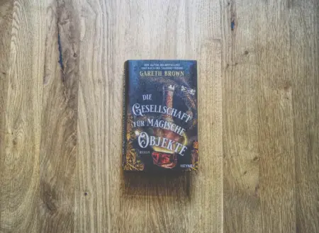 Ein deutsches Buch mit dem Titel "Die Gesellschaft für Magische Objekte" von Gareth Brown liegt auf einer Holzfläche.