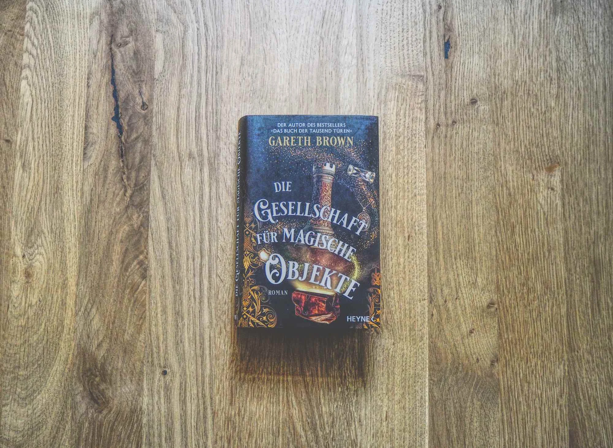 Ein deutsches Buch mit dem Titel "Die Gesellschaft für Magische Objekte" von Gareth Brown liegt auf einer Holzfläche.