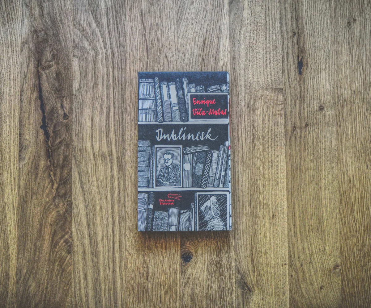 Auf einer Holzfläche liegt ein Buch mit dem Titel „Dublinesk“ von Enrique Vila-Matas. Auf dem Cover sind Bücherregale und ein lesender Mann abgebildet.