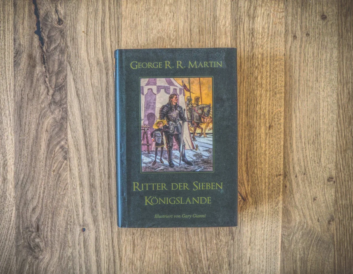Ein gebundenes Buch mit dem Titel „Ritter der Sieben Königslande“ von George R. R. Martin, mit einem illustrierten Cover mit mittelalterlichen Figuren, ruht auf einer Oberfläche mit Holzstruktur.