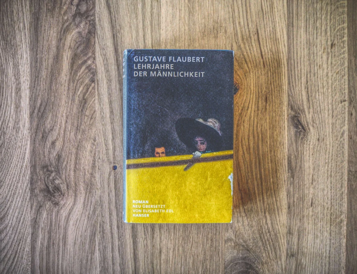 Eine deutsche Ausgabe von Gustave Flauberts Buch „Lehrjahre der Männlichkeit“ liegt auf einer Holzfläche. Auf dem Cover ist eine Frau abgebildet, die über einen Zaun lugt.