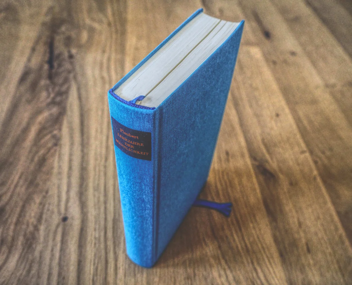 Ein gebundenes Buch mit blauem Einband und einem Lesebändchen steht aufrecht auf einer Holzfläche.