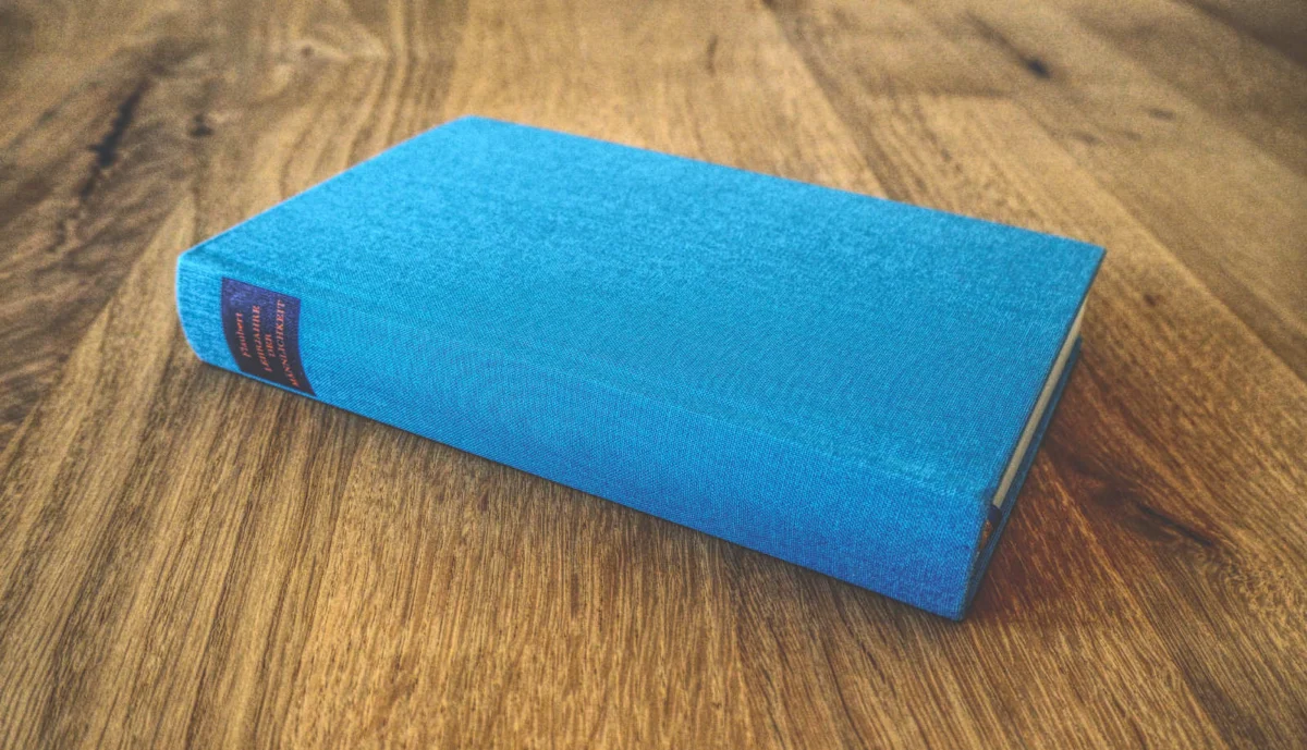 Ein geschlossenes Hardcover-Buch mit blauem Einband und rotem Textetikett auf dem Rücken liegt auf einer Holzoberfläche.