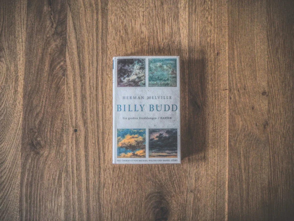 Auf einer Holzfläche liegt ein Buch mit dem Titel „Billy Budd“ von Herman Melville. Der Einband ist mit einem Mosaik aus kleinen Bildern verziert.