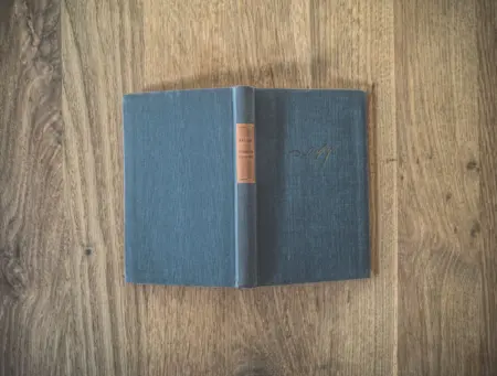 Ein altes gebundenes Buch mit grünem Einband und braunem Buchrücken und dunklem Text liegt aufgeschlagen auf einer Holzoberfläche. Der Titel des Buches ist nicht zu sehen.