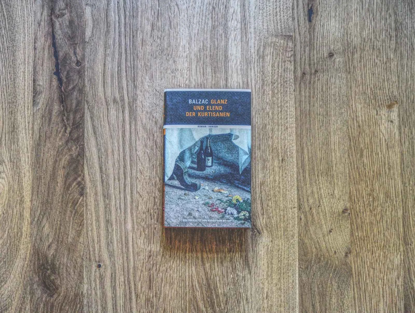 Ein Buch mit dem Titel "Balzac Glanz und Elend der Kurtisanen" liegt geschlossen auf einer Holzplatte.