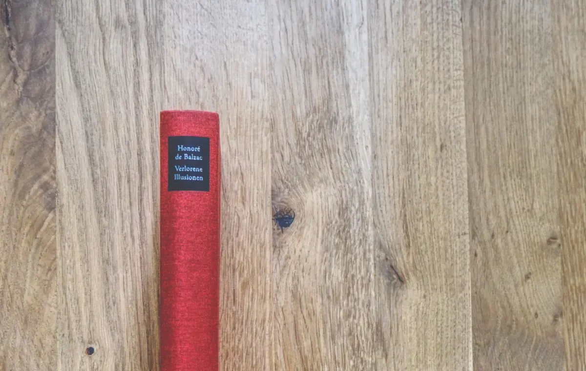 Ein rotes Buch mit dem Titel "Honoré de Balzac, Verlorene Illusionen" steht aufrecht auf einer Holzplatte.