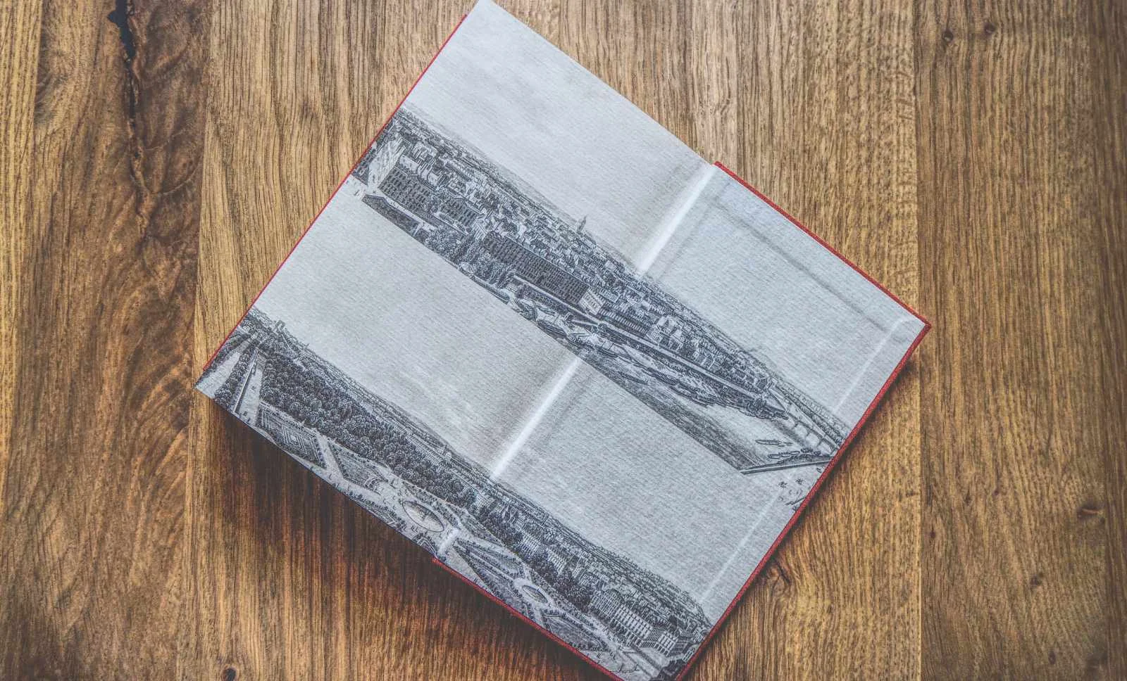 Ein aufgeschlagenes Buch auf einer hölzernen Oberfläche, das auf beiden Seiten eine Schwarz-Weiß-Illustration einer Stadt zeigt.