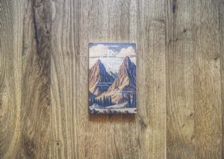 Auf einer Holzfläche liegt ein Buch mit dem Titel „Die Schwere Not“ mit einer Bergillustration auf dem Cover.