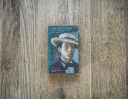 Auf einer Holzfläche liegt das Buch „Eine Gewöhnliche Geschichte“ von Iwan Gontscharow. Auf dem Cover ist das Porträt eines Mannes mit Hut und Anzug zu sehen.