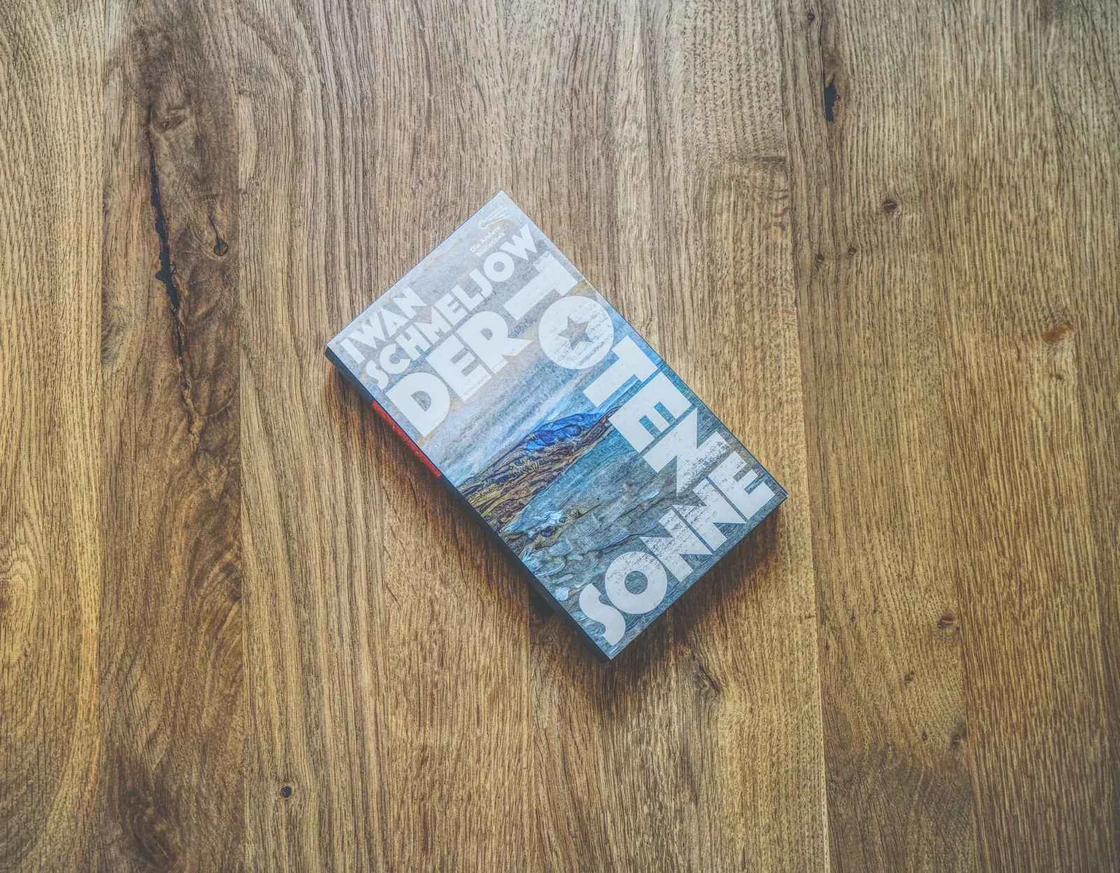 Ein Buch mit dem Titel "Der Toten Sonne" von Iwan Schmeljow liegt auf einer Holzfläche. Der Einband zeigt ein Landschaftsbild mit großer weißer Schrift.