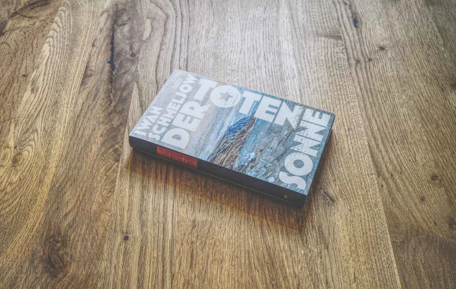 Ein Buch mit dem Titel "Der Toten Sonne" von Iwan Schmeljow liegt auf einer Holzfläche. Der Einband zeigt ein graues und braunes abstraktes Muster mit großer weißer Schrift.