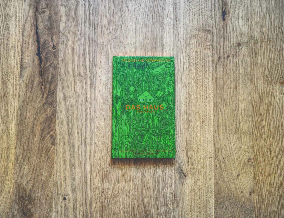 Ein grünes Buch mit dem Titel "Das Haus" liegt mittig auf einer hellen Holzfläche.