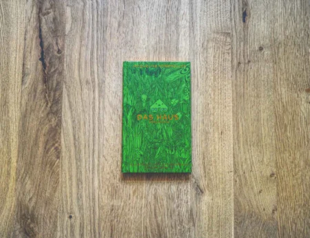 Ein grünes Buch mit dem Titel "Das Haus" liegt mittig auf einer hellen Holzfläche.