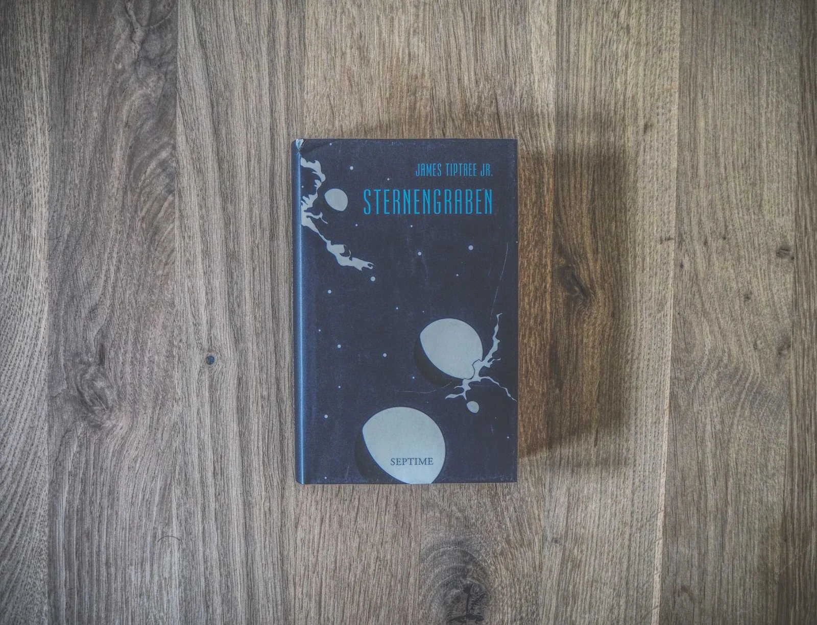 Ein Buch mit dem Titel „Sternengraben“ von James Tiptree Jr. liegt zentriert auf einer Holzoberfläche. Der Einband ist schwarz mit weißen abstrakten Mustern und blauem Text.