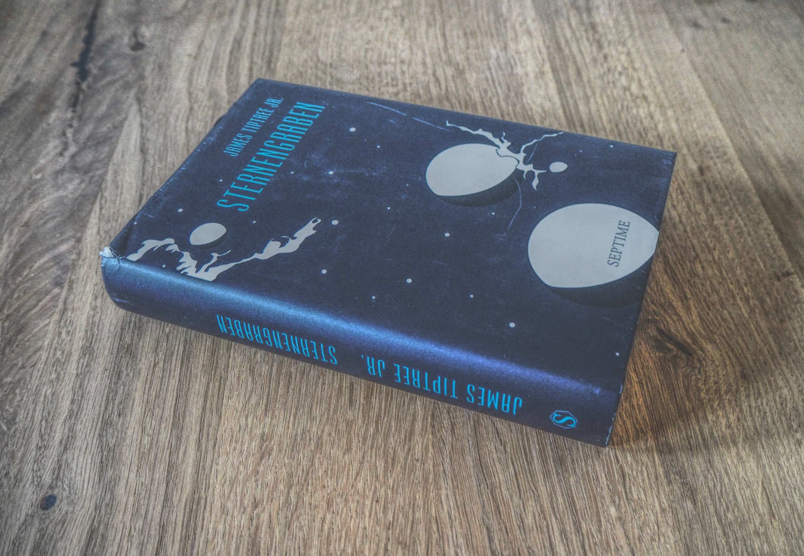 Ein gebundenes Buch mit dem Titel „Sternengraben“ von James Tiptree Jr. mit einem dunkelblauen Cover, auf dem zwei Planeten und ein schwebender Astronaut abgebildet sind. Das Buch liegt auf einer Holzoberfläche.