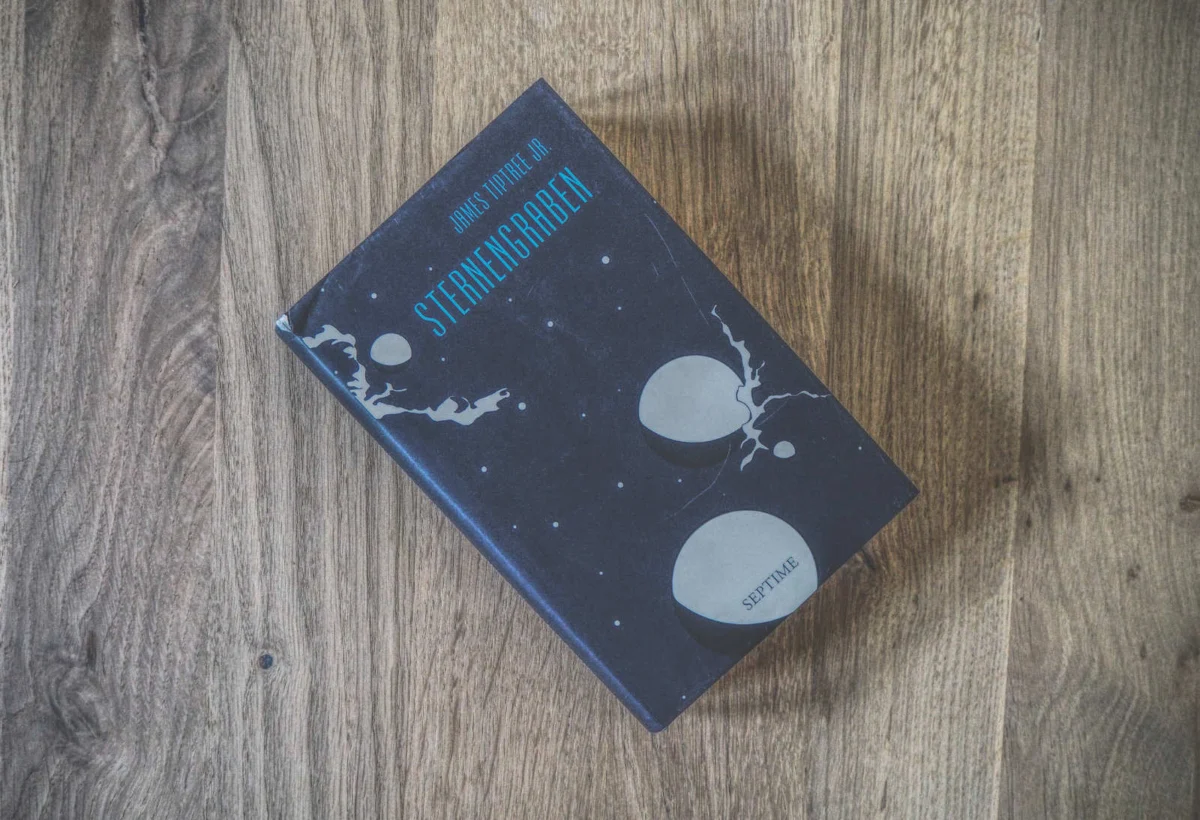 Auf einer Holzfläche liegt ein Buch mit dem Titel „Sternengraben“ von James Tiptree Jr., dessen dunkler Einband mit abstrakten, kosmischen Illustrationen verziert ist.