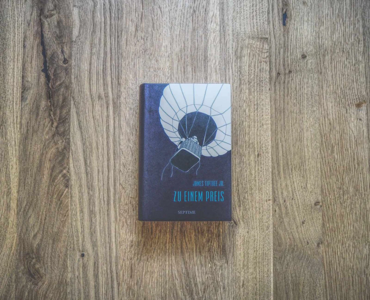 Auf einer Holzfläche liegt ein Buch mit dem Titel „Zu einem Preis“ von James Tiptree Jr. mit dem Coverbild eines Heißluftballons.
