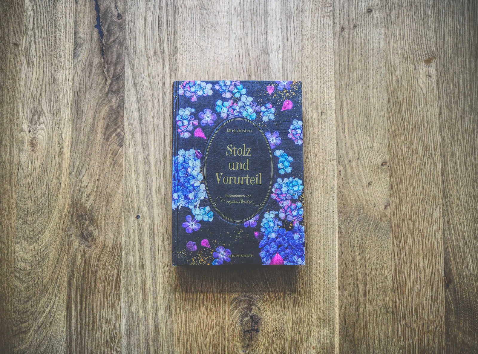 Auf einer Holzfläche liegt ein Buch mit dem Titel „Stolz und Vorurteil“ von Jane Austen. Auf dem Einband sind violette und blaue Blumen abgebildet.