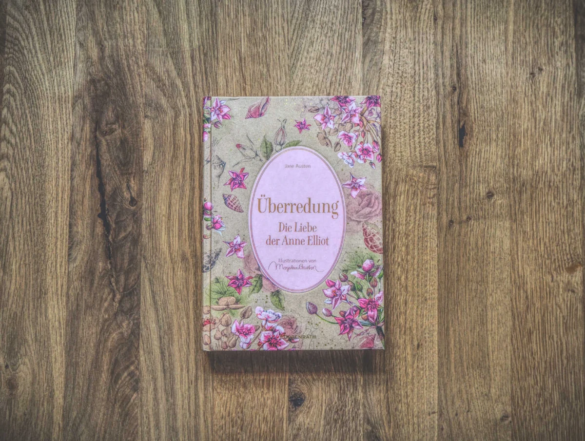 Auf einer Holzfläche liegt eine deutsche Ausgabe des Buches „Überredung“ von Jane Austen mit floralen Motiven auf dem Cover und dem Titel „Die Liebe der Anne Elliot“.