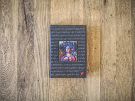 Auf einer Holzoberfläche liegt ein Buch mit dunklem Einband, in dessen Mitte die Abbildung einer Frau, umgeben von Text, zu sehen ist.