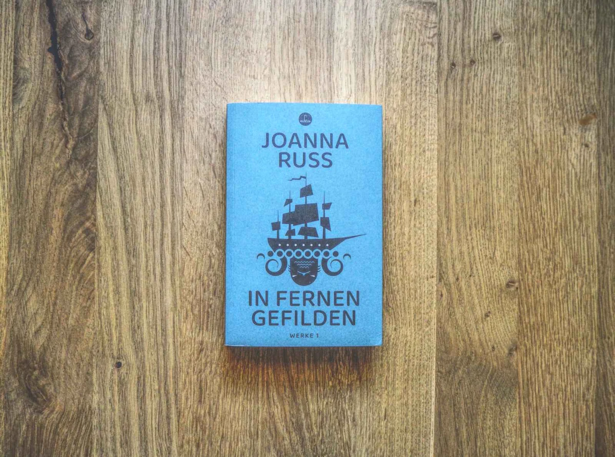Ein blaues Buch mit dem Titel „Joanna Russ In Fernen Gefilden“ mit einer Schiffsillustration auf dem Cover, platziert auf einer Holzoberfläche.