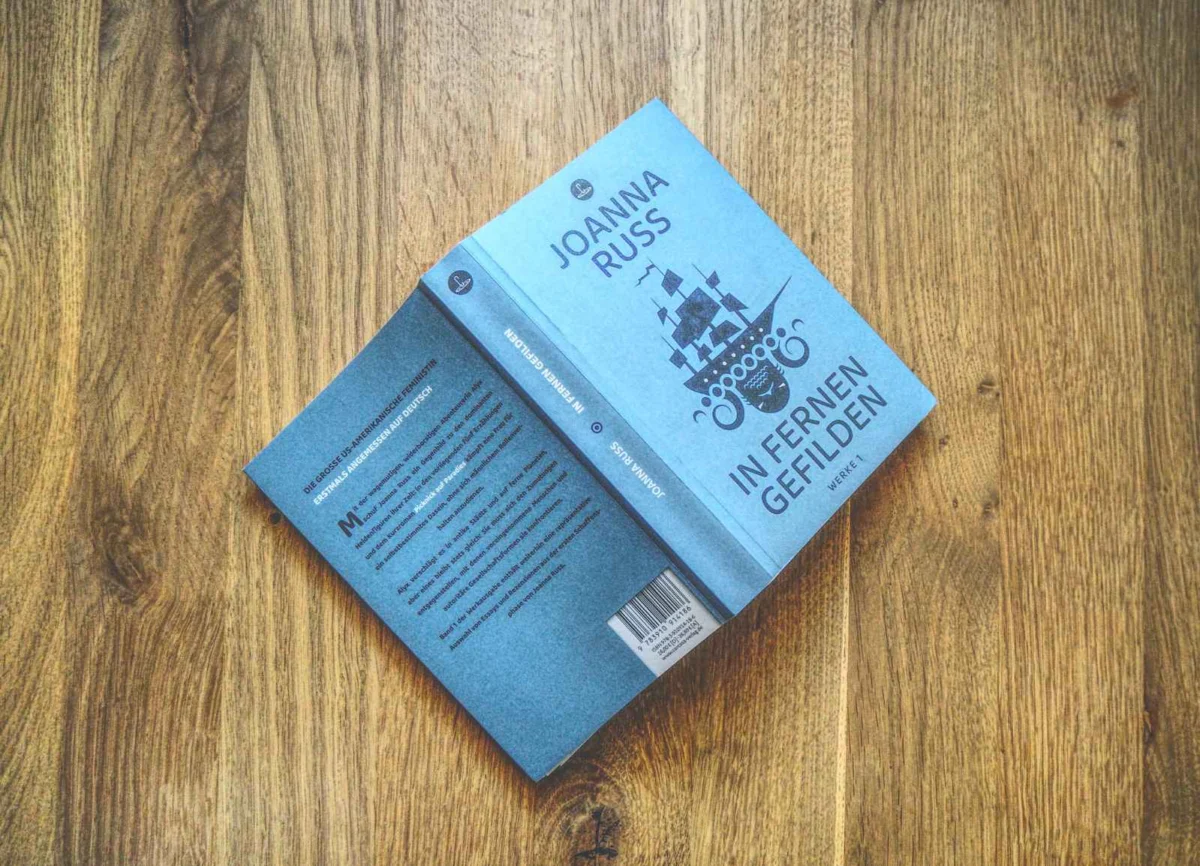 Ein blaues Buch mit dem Titel „Joanna Russ: In Fernen Gefilden“ liegt aufgeschlagen auf einer Holzoberfläche. Zu sehen sind der vordere Einband, der Buchrücken und ein Teil des hinteren Einbands.