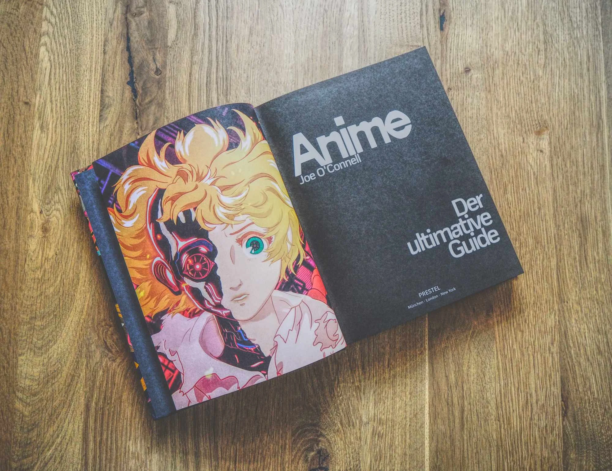 Ein aufgeschlagenes Buch auf einer Holzfläche zeigt auf der linken Seite eine illustrierte Anime-Figur und auf der rechten Seite den Titel "Anime: Der ultimative Guide" von Joe O'Connell auf der rechten Seite.