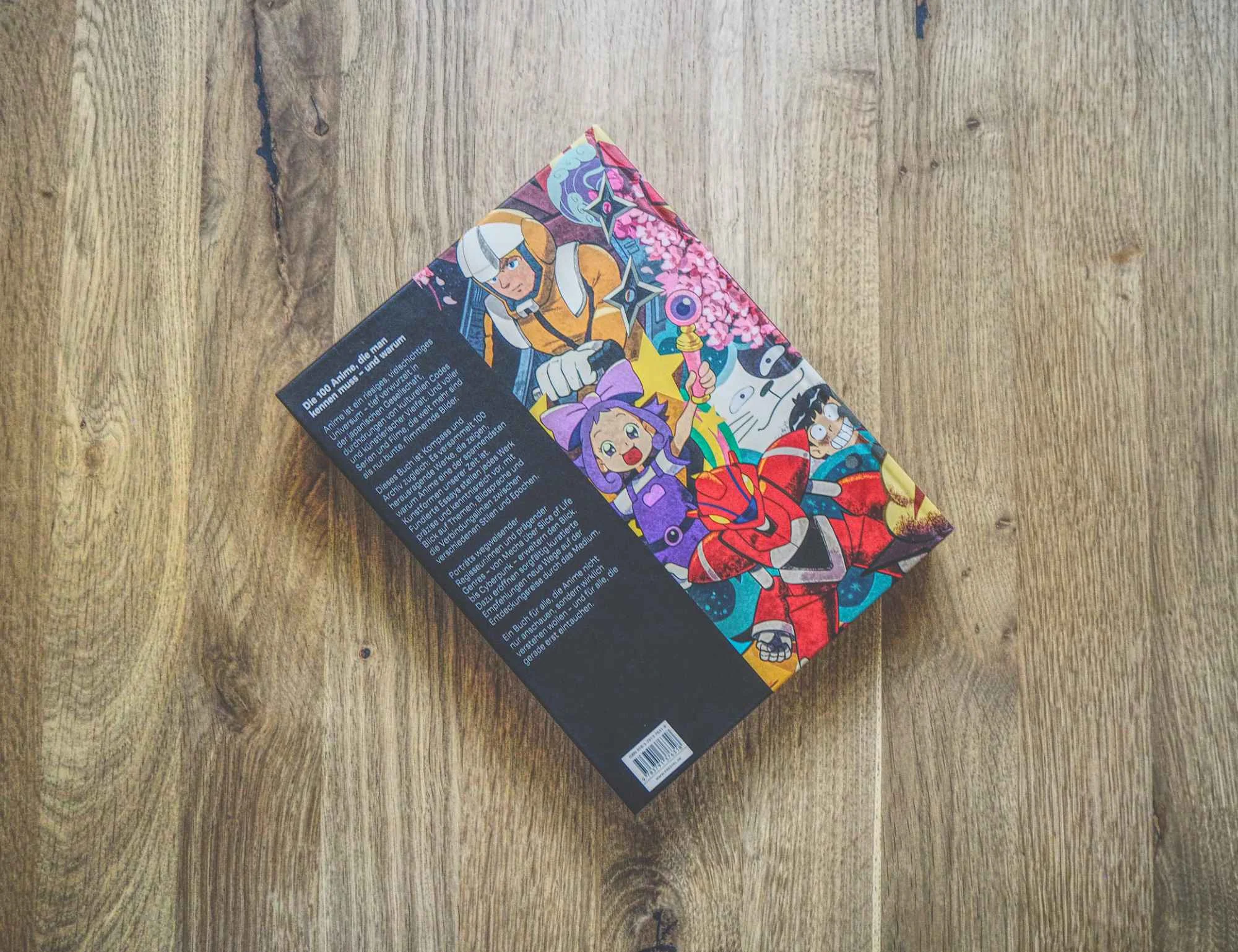 Ein Buch mit einem farbenfroh illustrierten Einband, auf dem animierte Figuren zu sehen sind, liegt mit dem Rücken nach unten auf einer hölzernen Unterlage und zeigt seinen Einband und den Strichcode.