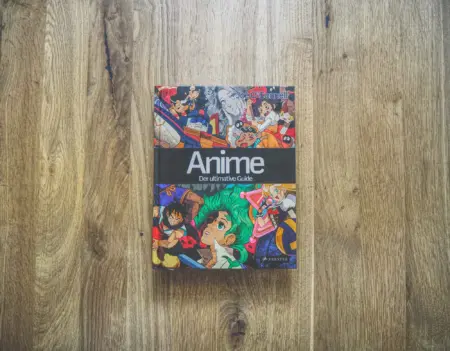 Ein Hardcover-Buch mit dem Titel "Anime: Der ultimative Guide" mit einem farbenfrohen, illustrierten Einband, auf dem verschiedene Anime-Figuren abgebildet sind, steht auf einer Holzfläche.