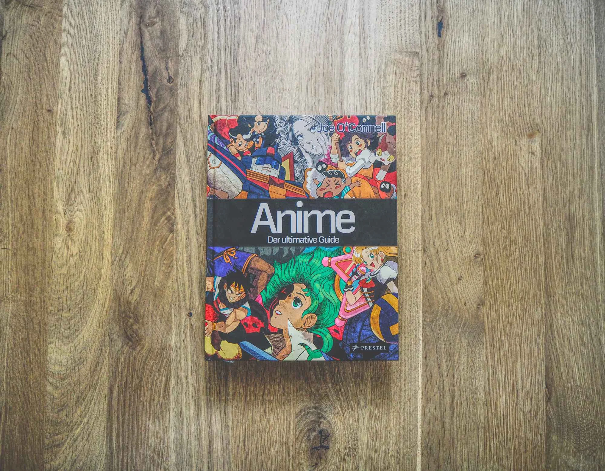 Ein Hardcover-Buch mit dem Titel "Anime: Der ultimative Guide" mit einem farbenfrohen, illustrierten Einband, auf dem verschiedene Anime-Figuren abgebildet sind, steht auf einer Holzfläche.