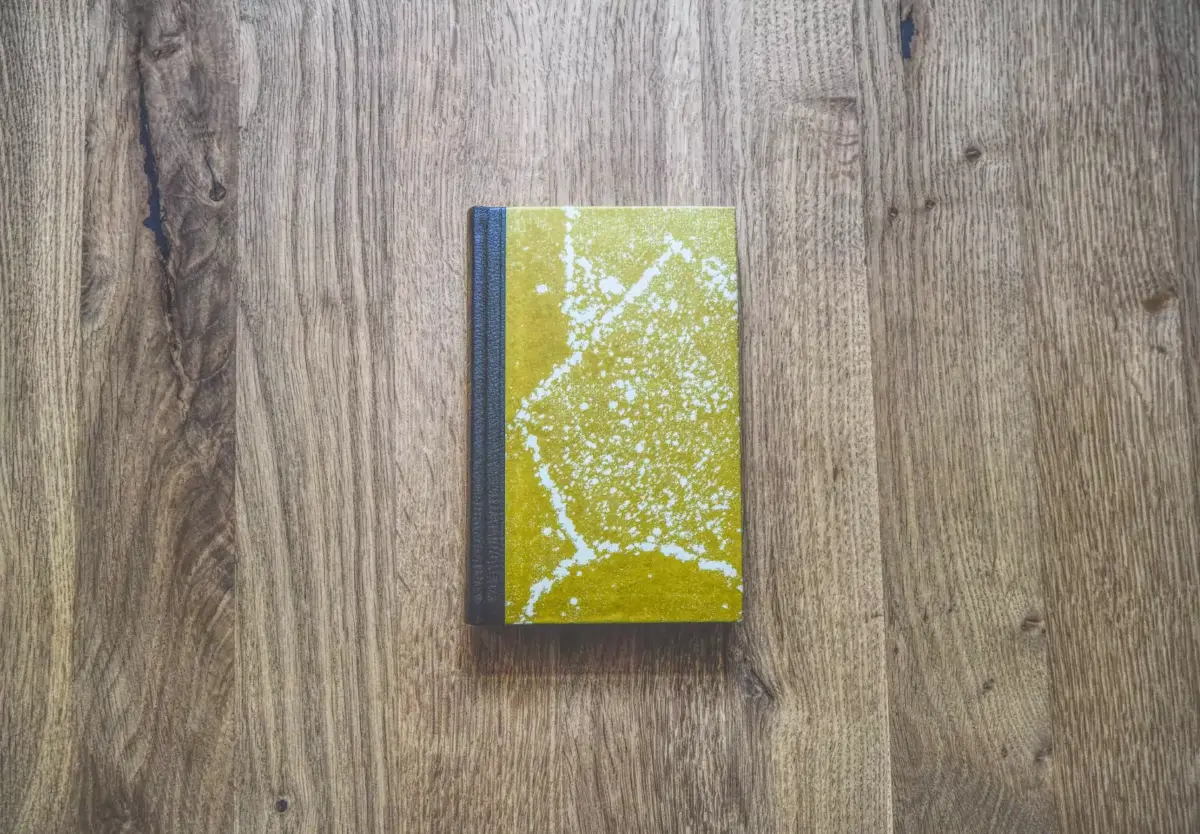 Ein kleines gelbes Buch mit schwarzem Buchrücken liegt geschlossen auf einer Holzoberfläche. Der Buchdeckel weist ein marmoriertes Muster auf.