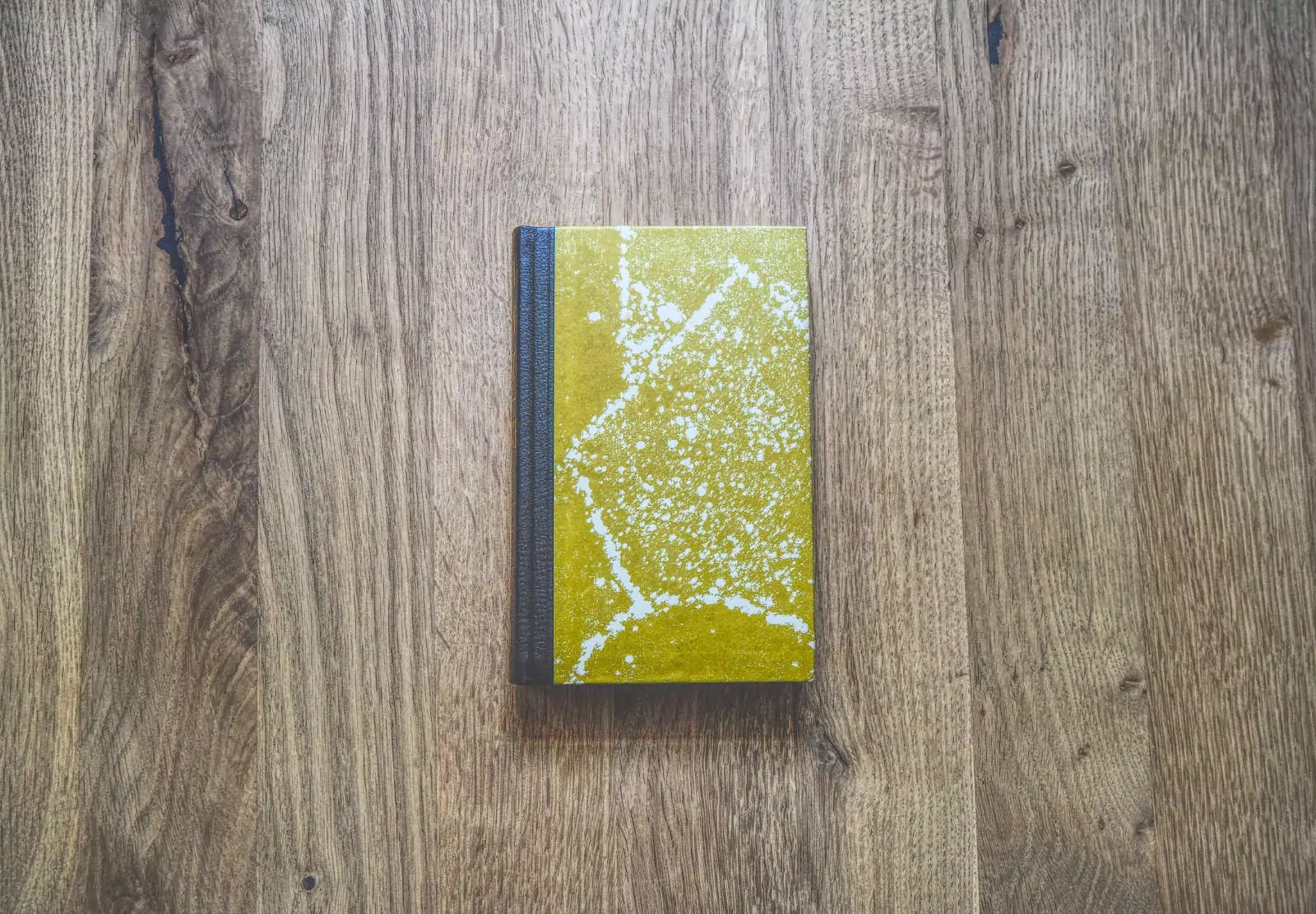 Ein kleines gelbes Buch mit schwarzem Buchrücken liegt geschlossen auf einer Holzoberfläche. Der Buchdeckel weist ein marmoriertes Muster auf.