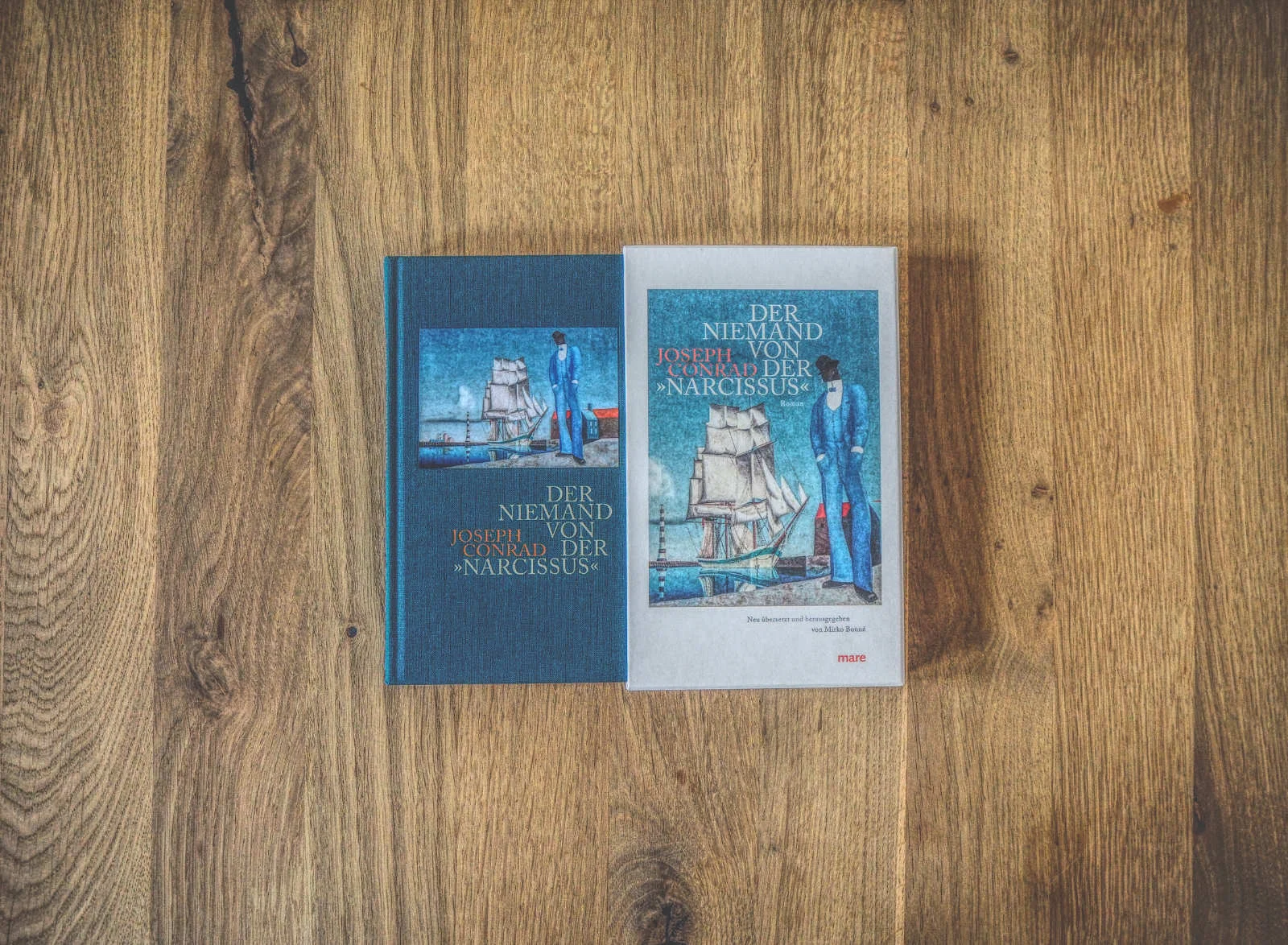 Zwei Bücher mit dem Titel „Der Niemand von der Narcissus“ von Joseph Conrad, die ein Schiff und einen Mann abbilden, liegen nebeneinander auf einer Holzfläche.