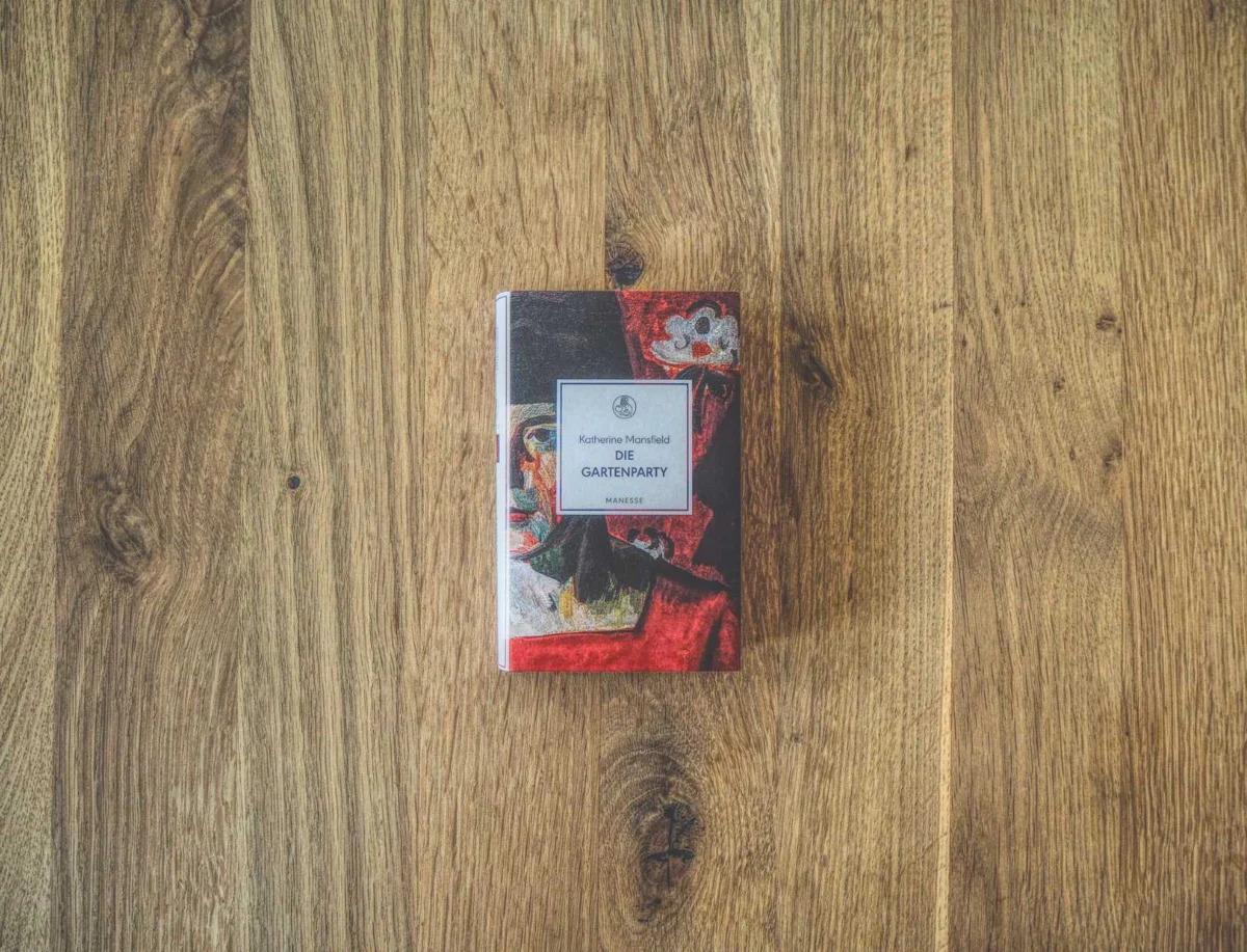 Ein kleines Buch mit dem Titel „Die Gartenparty“ von Katherine Mansfield, dessen Cover ein abstraktes Gemälde auf einer Holzoberfläche zeigt.