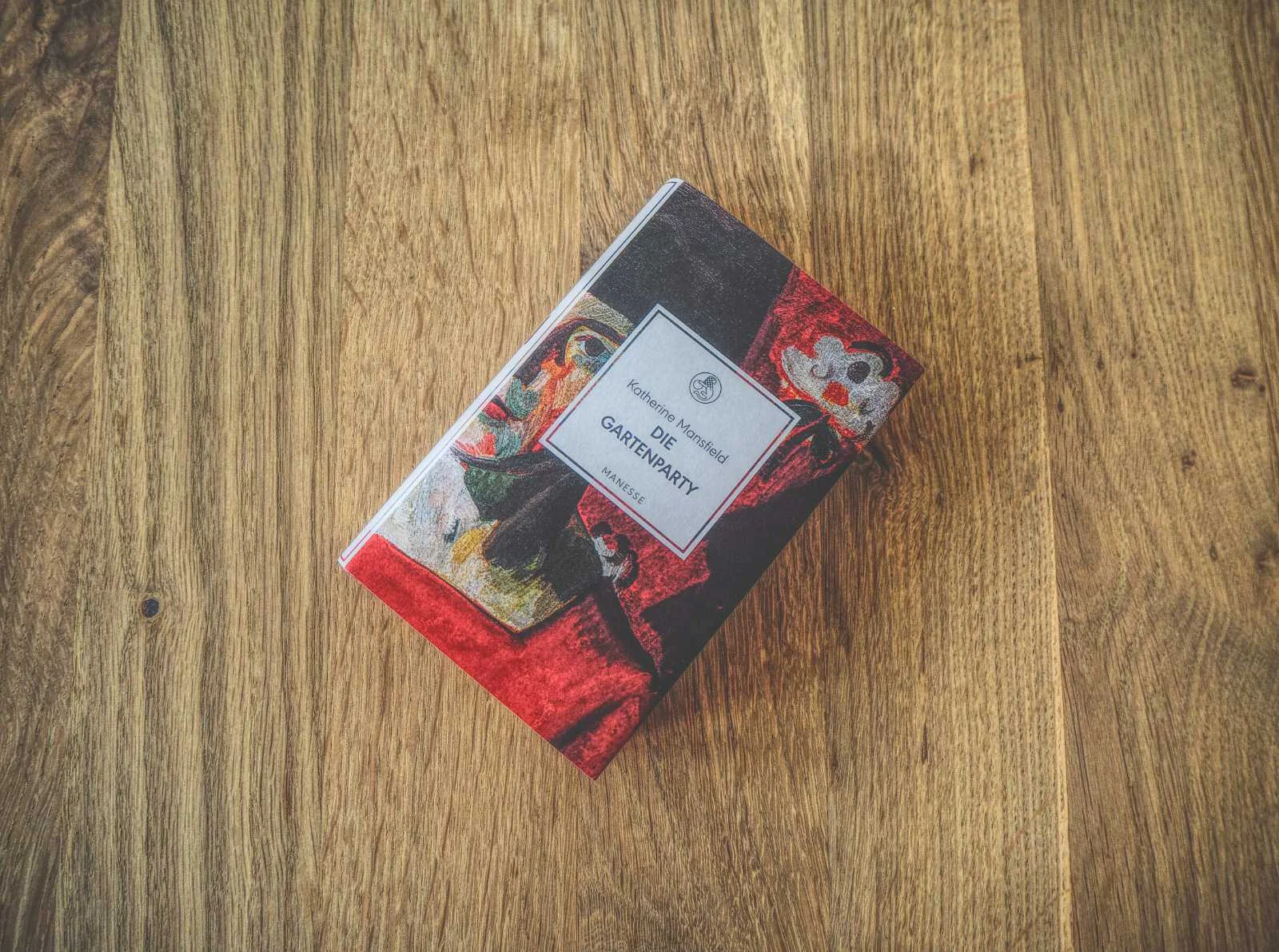 Auf einer Holzoberfläche liegt ein Buch mit dem Titel „Die Gartenparty“ von Katherine Mansfield mit einem farbenfrohen abstrakten Cover.