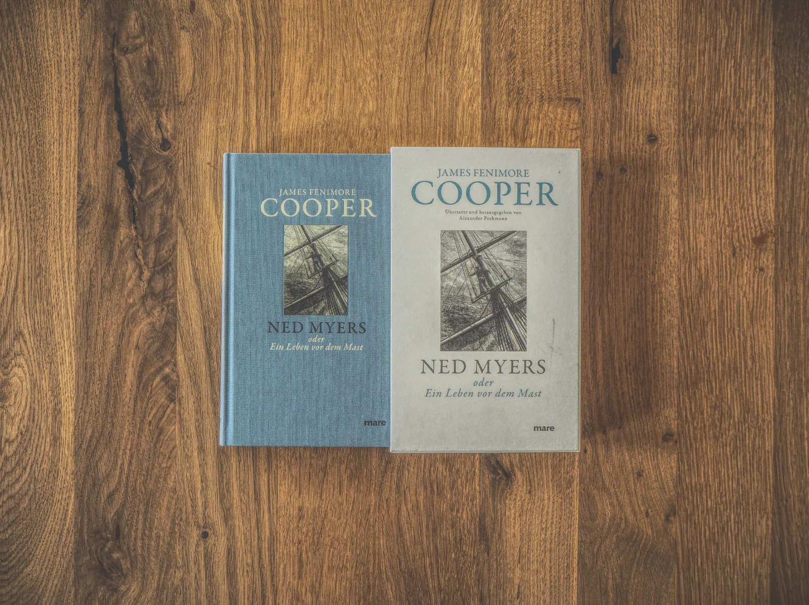 Zwei Bücher mit dem Titel „Ned Myers“ von James Fenimore Cooper liegen auf einer Holzoberfläche. Ein Buch hat einen blauen und das andere einen grauen Einband. Beide zeigen das gleiche Cover.