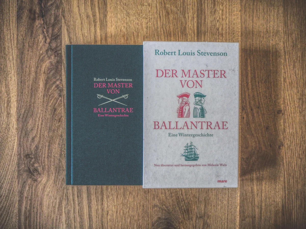 Auf einer Holzoberfläche liegen zwei Bücher mit dem Titel „Der Master von Ballantrae“ von Robert Louis Stevenson. Ein Buch hat einen dunklen Einband, das andere ist heller und zeigt Abbildungen von zwei Männern und einem Schiff.