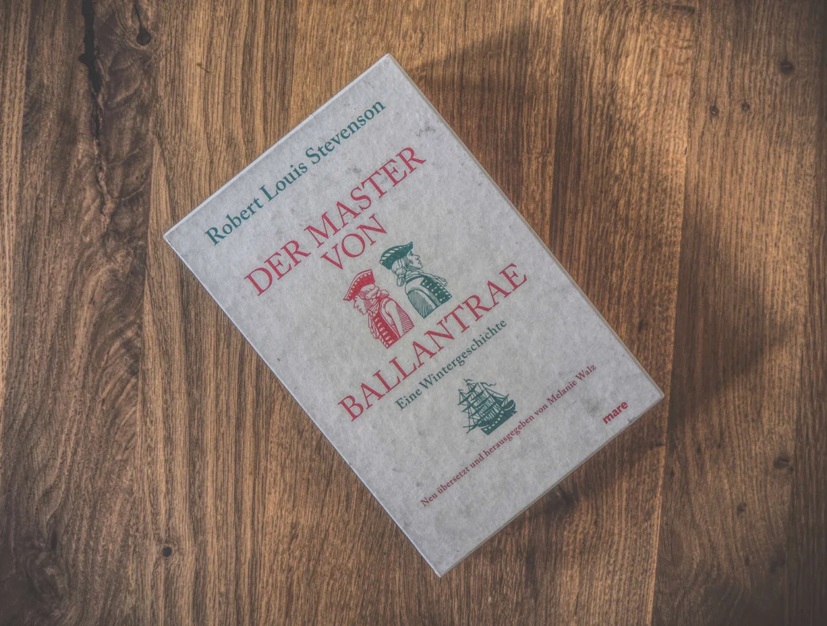 Auf einer Holzfläche liegt ein Buch mit dem Titel „Der Master von Ballantrae“ von Robert Louis Stevenson.