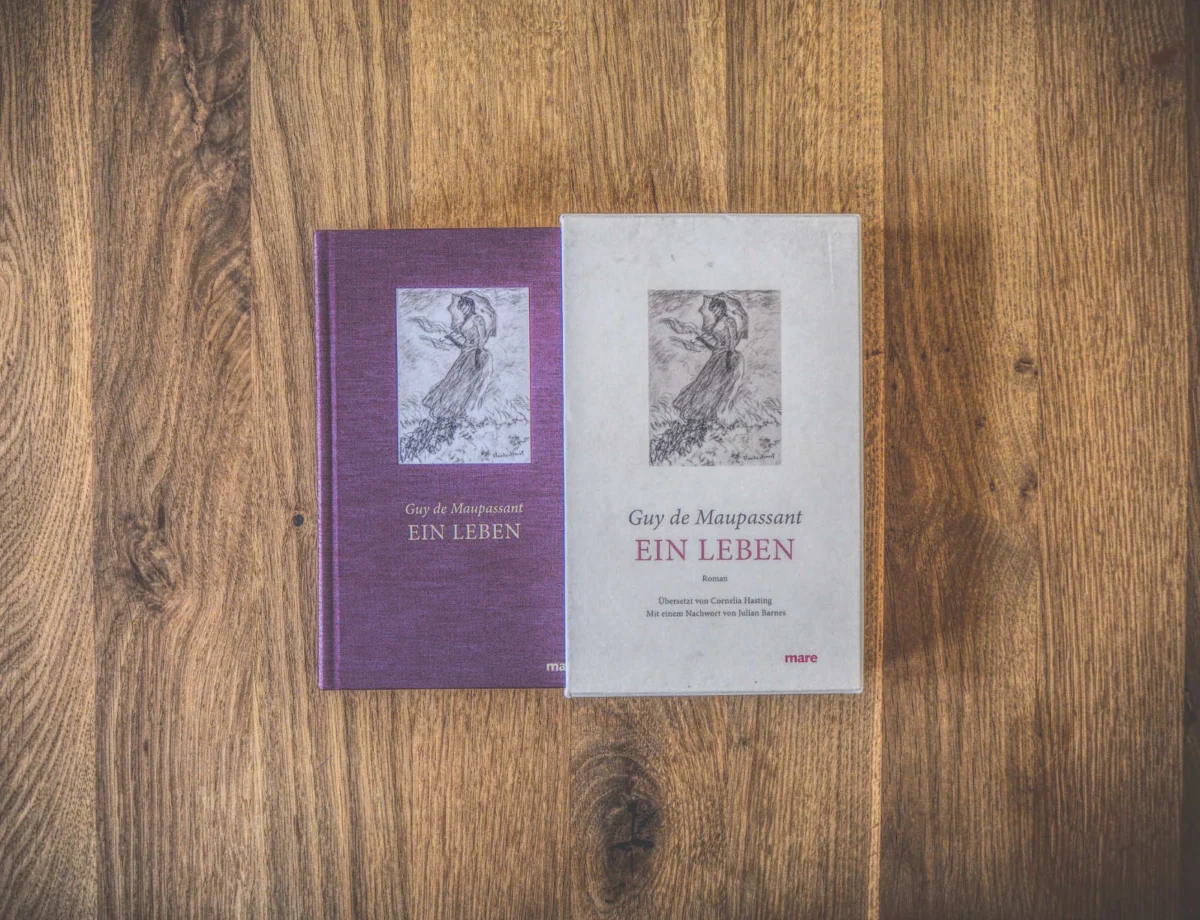 Zwei Exemplare von Guy de Maupassants Buch „Ein Leben“ liegen nebeneinander auf einer Holzfläche. Das linke Exemplar hat einen violetten Einband, das rechte Exemplar einen beigen.