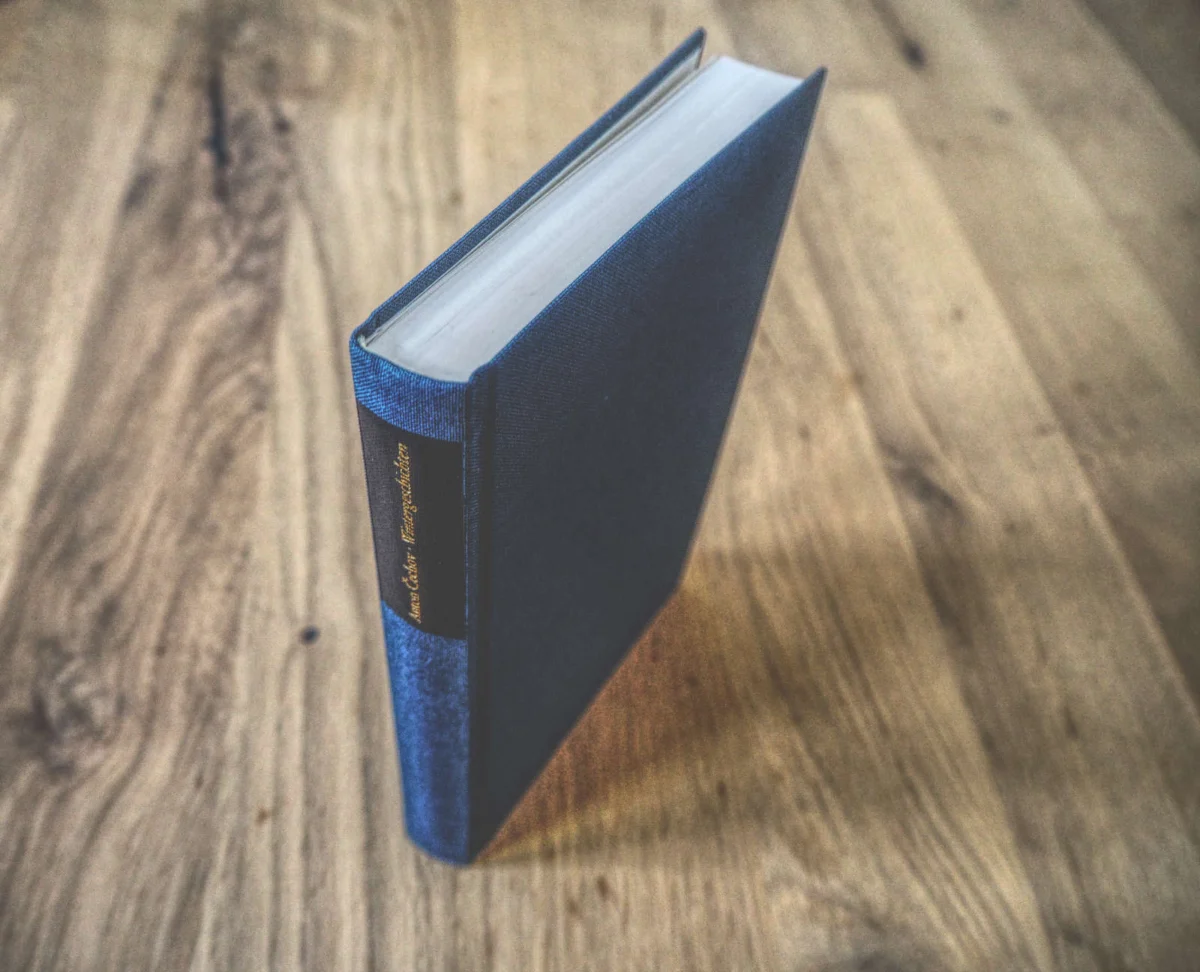 Auf einer Holzfläche steht ein Hardcover-Buch mit blauem Einband. Der Buchrücken ist mit goldenem Text versehen.