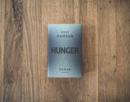 Auf einer Holzfläche liegt ein Buch mit dem Titel „Hunger“ von Knut Hamsun. Der Einband ist grau mit schwarzem Text.