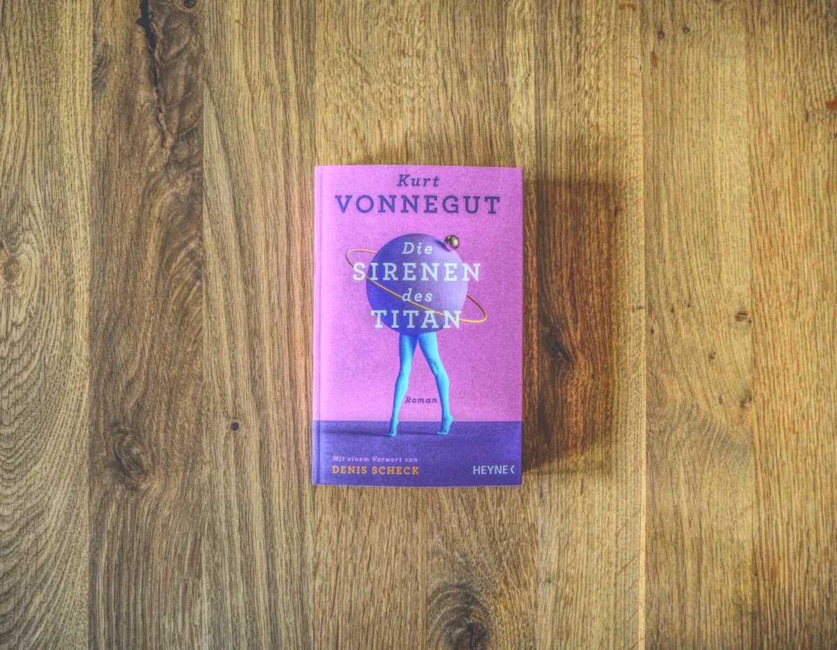 Ein Buch mit dem Titel „Die Sirenen des Titan“ von Kurt Vonnegut, mit einem violetten Einband und der Abbildung einer Person und eines Planeten mit Ringen, liegt auf einer Holzoberfläche.