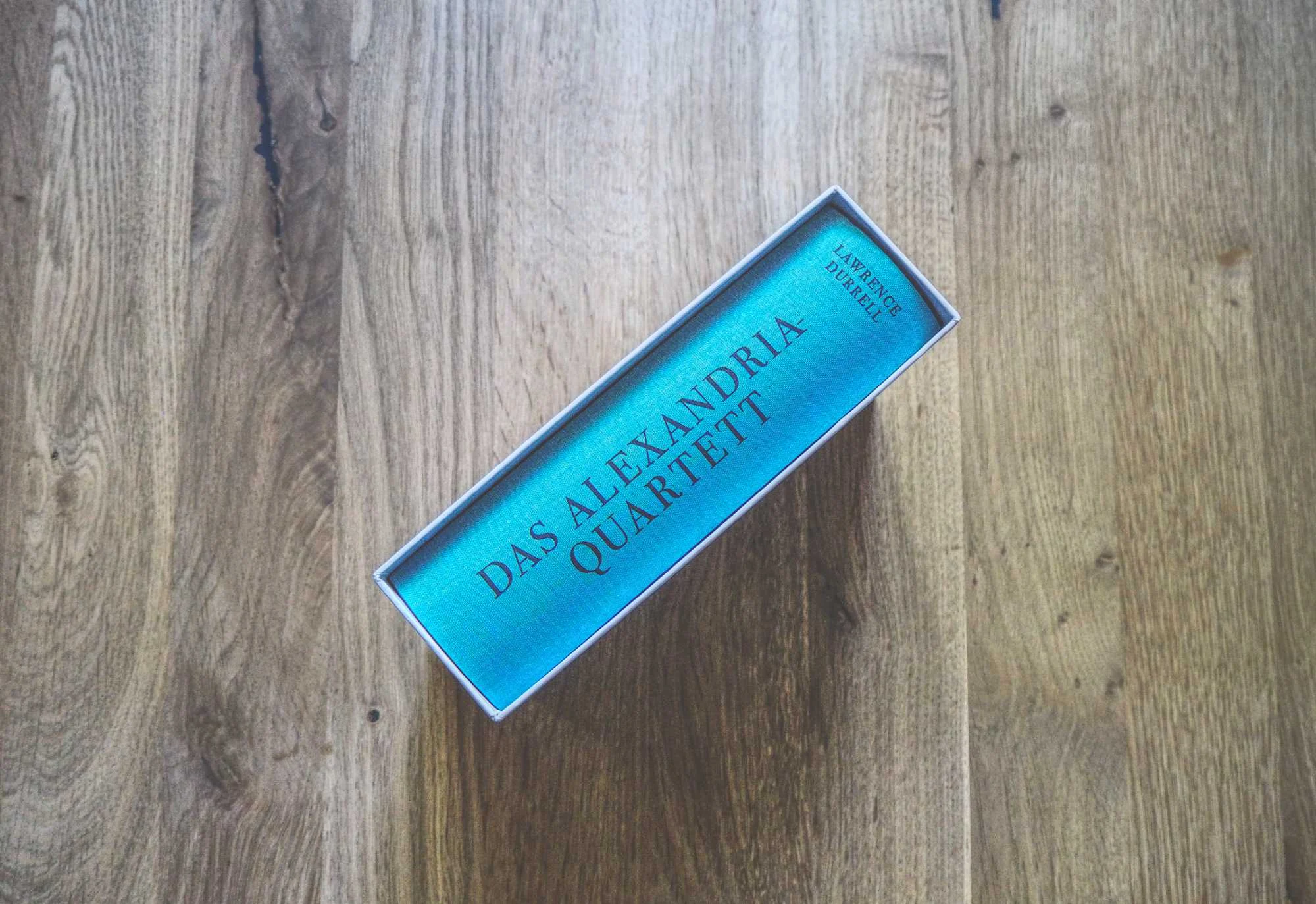 Ein blaues Buch mit dem Titel "Das Alexandria Quartett" von Lawrence Durrell wird zugeklappt und auf eine Holzfläche gelegt.