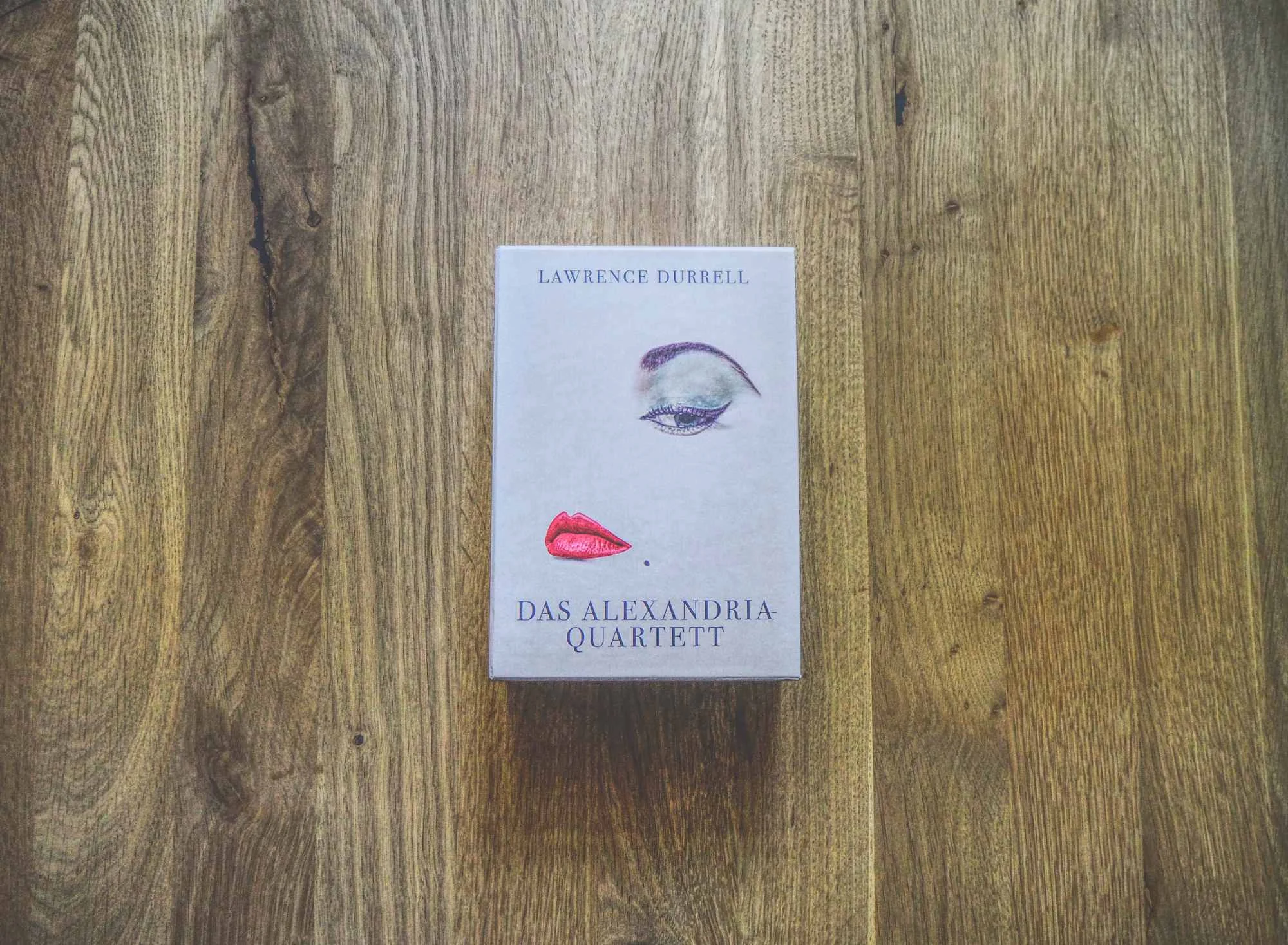 Auf einer hölzernen Unterlage liegt eine Box mit "Das Alexandria Quartett" von Lawrence Durrell; das Cover zeigt ein Auge und ein Paar rote Lippen.