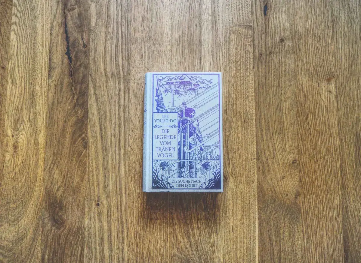 Ein gebundenes Buch mit dem Titel "Die Legende von Tränenvogel" von Lee Young-Do liegt geschlossen auf einer Holzfläche.