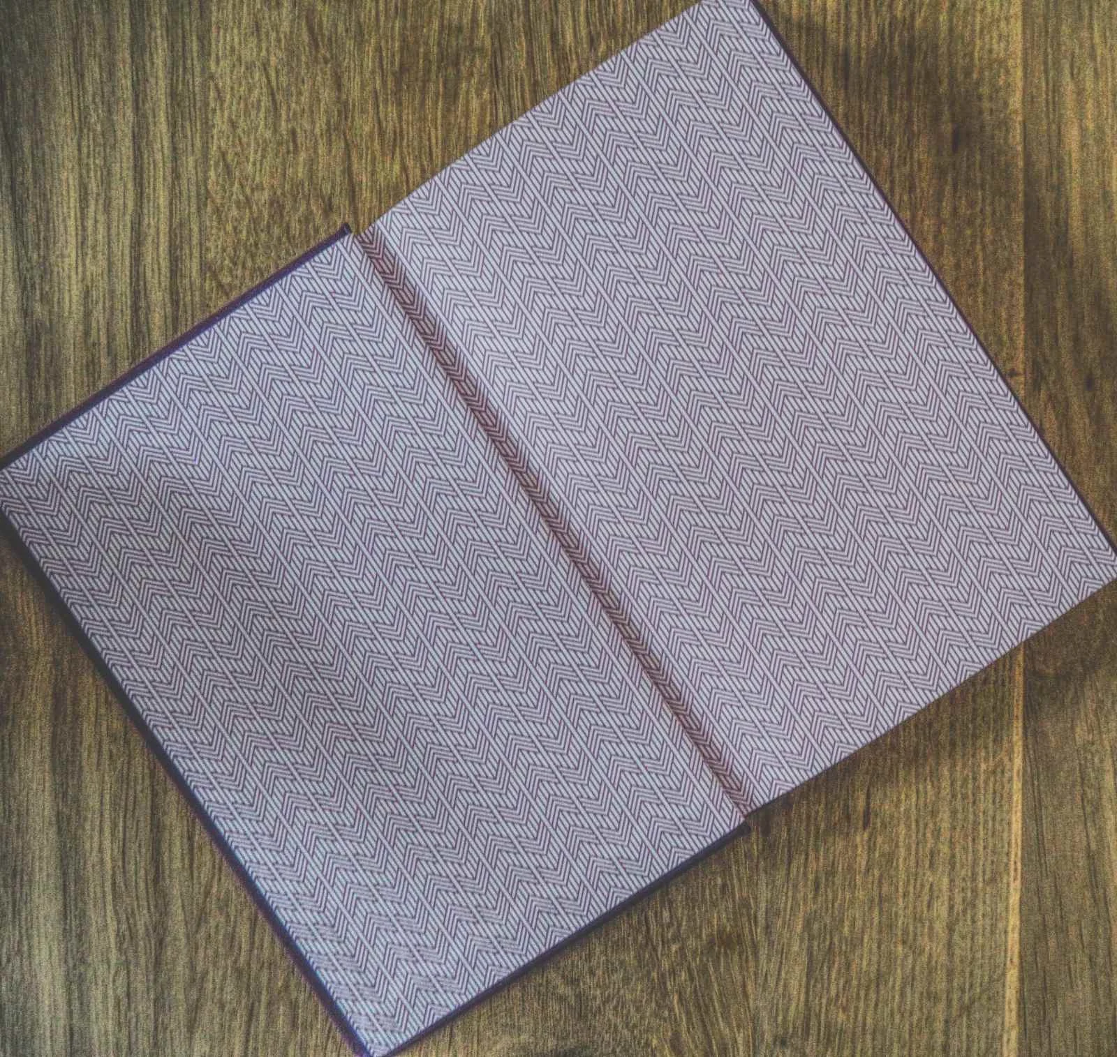 Ein offenes Buch liegt flach auf einer Holzoberfläche und zeigt gemusterte, rosafarbene Vorsatzblätter mit einem sich wiederholenden Winkelmuster.