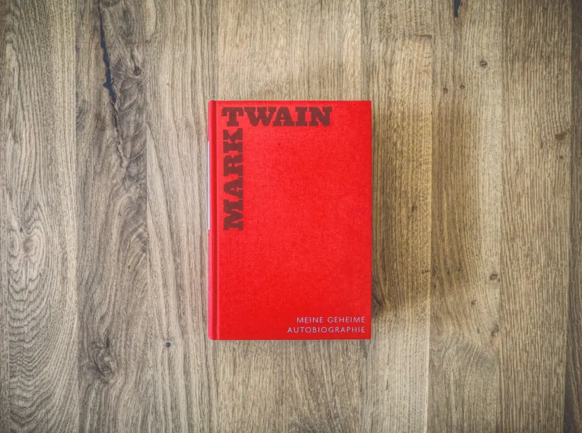 Auf einer Holzunterlage liegt ein rotes Buch mit dem Titel „Mark Twain: Meine geheime Autobiographie“.